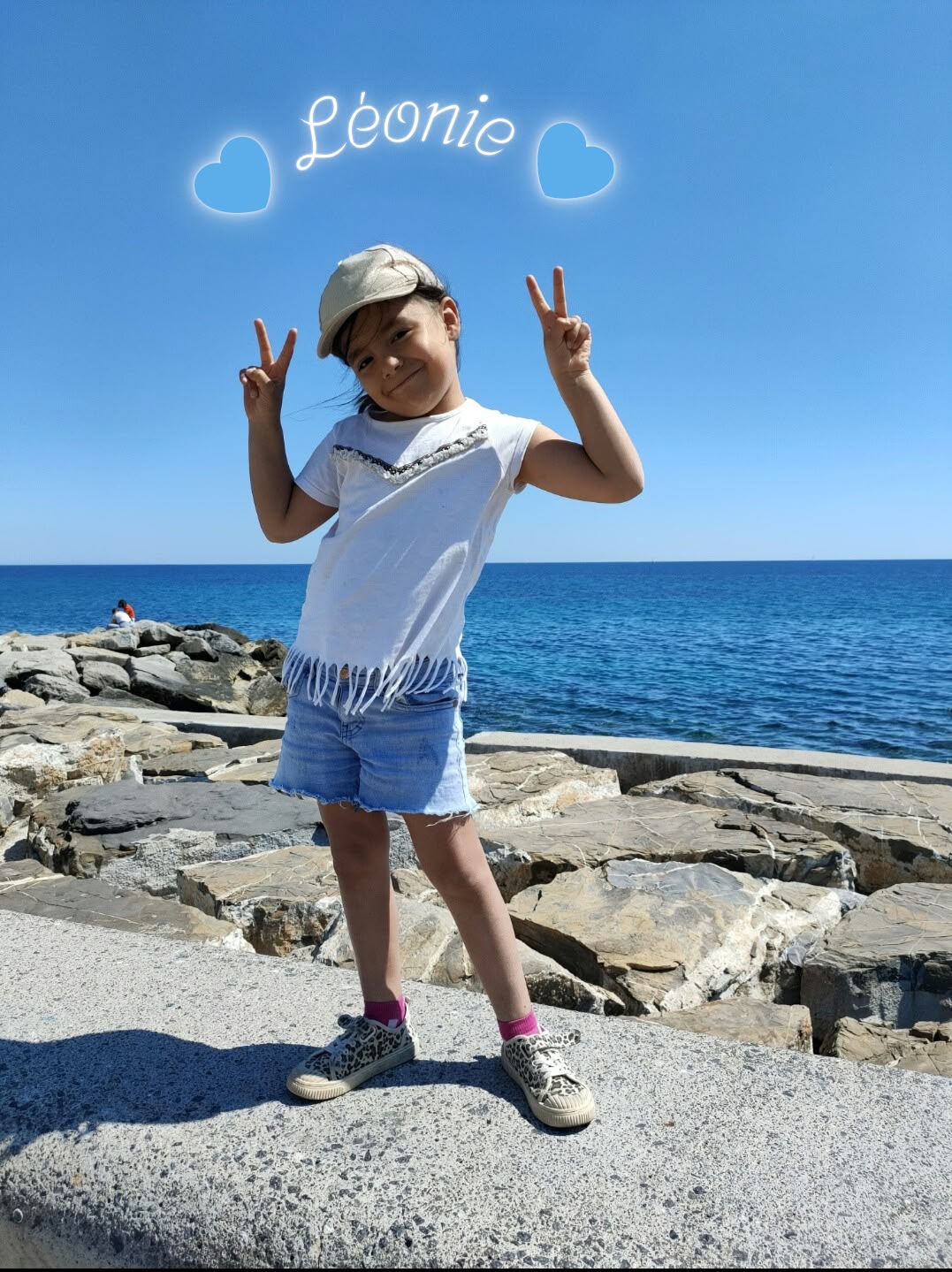 Léonie souriante au bord de la mer, faisant le signe de la victoire des deux mains